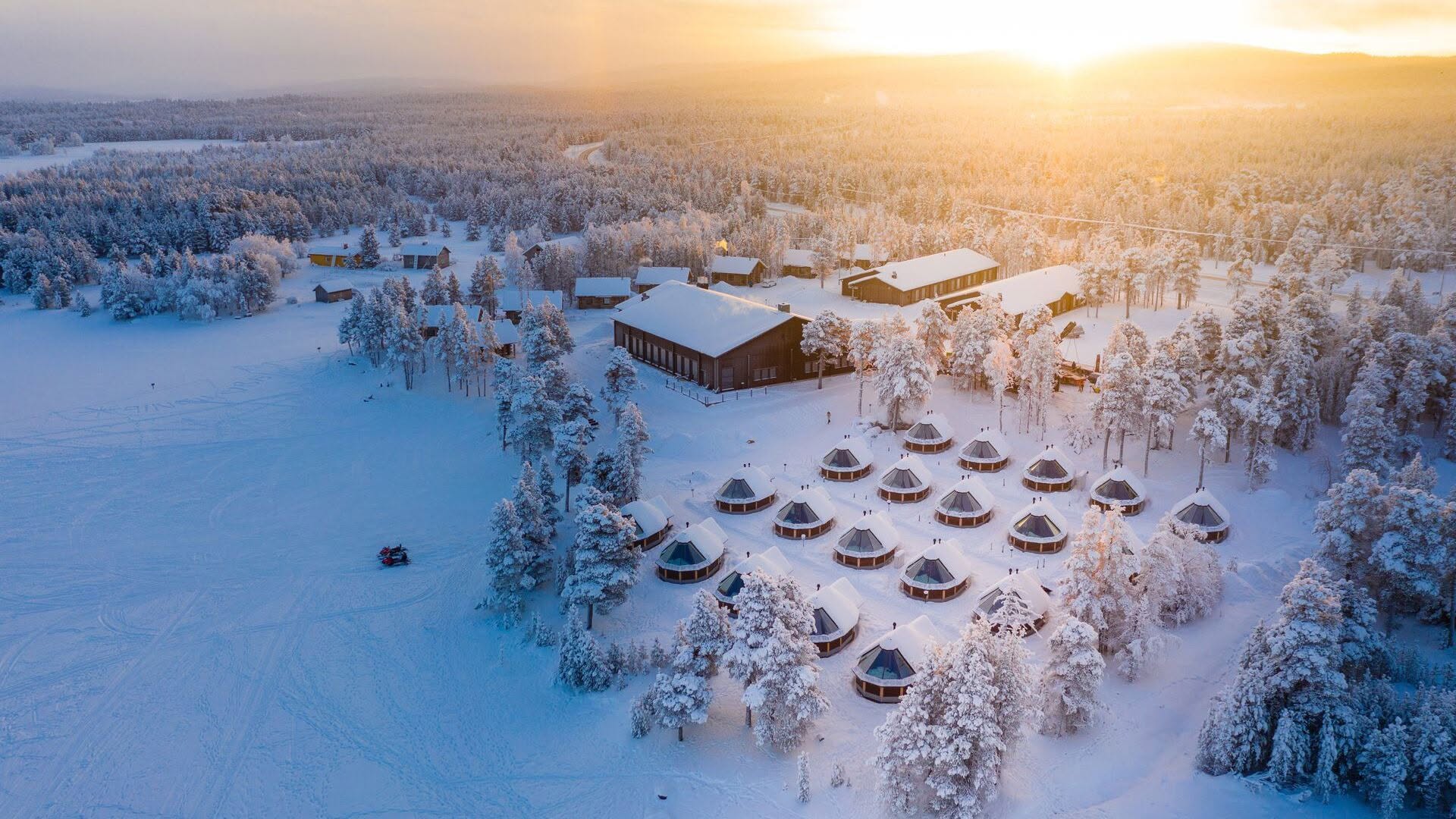 Aerial view of glass igloos and wooden lodge buildings at Wilderness Hotel Muotka & Nellim in snow-covered pine woods