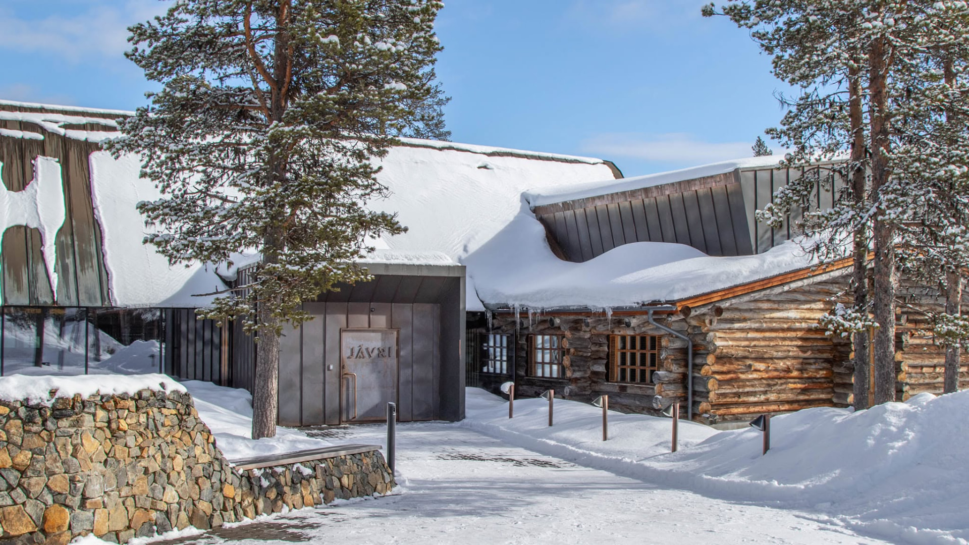 Snow-covered entrance to Jávri Lodge in Saariselkä with timber log walls and a dark metal façade