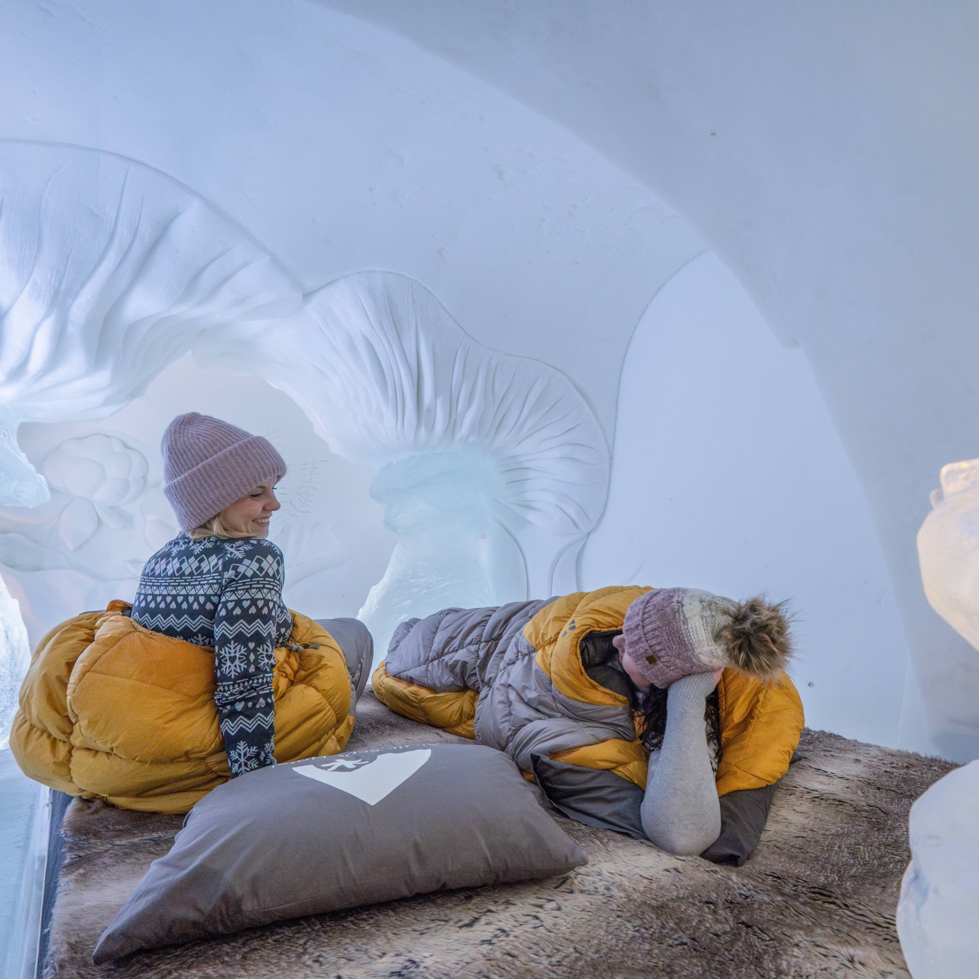 Guests in sleeping bags on a fur-topped bed inside a carved snow room at Arctic SnowHotel & Glass Igloos in Sinettä, Rovaniemi